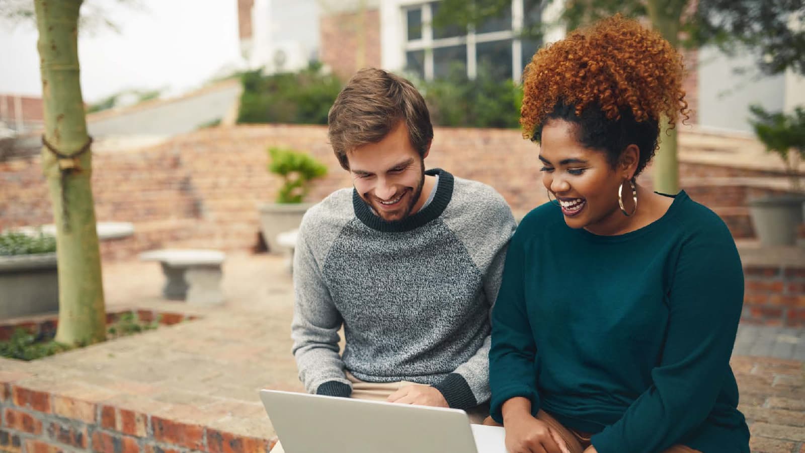 2 students laugh while looking at a laptop together on a college campus